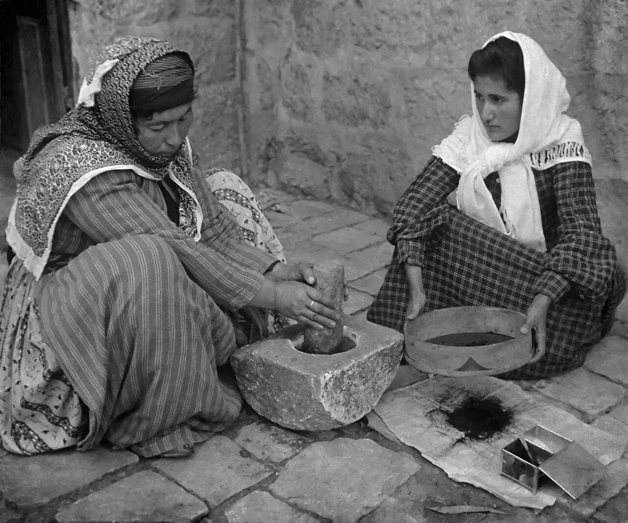 Two women grinding coffee beans by hand with a stone mortar in an old photograph