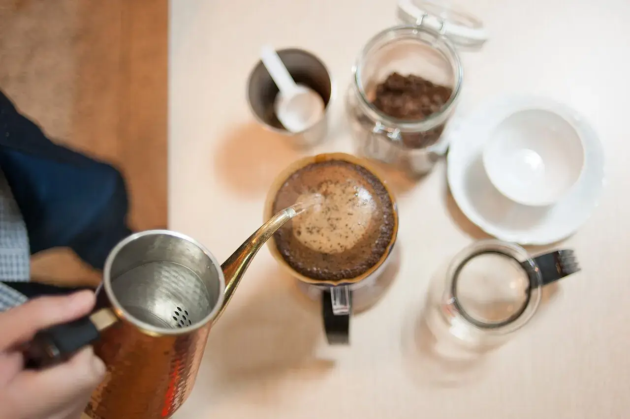 Hot water being poured into a brewer, with a jar of coffee and cups nearby